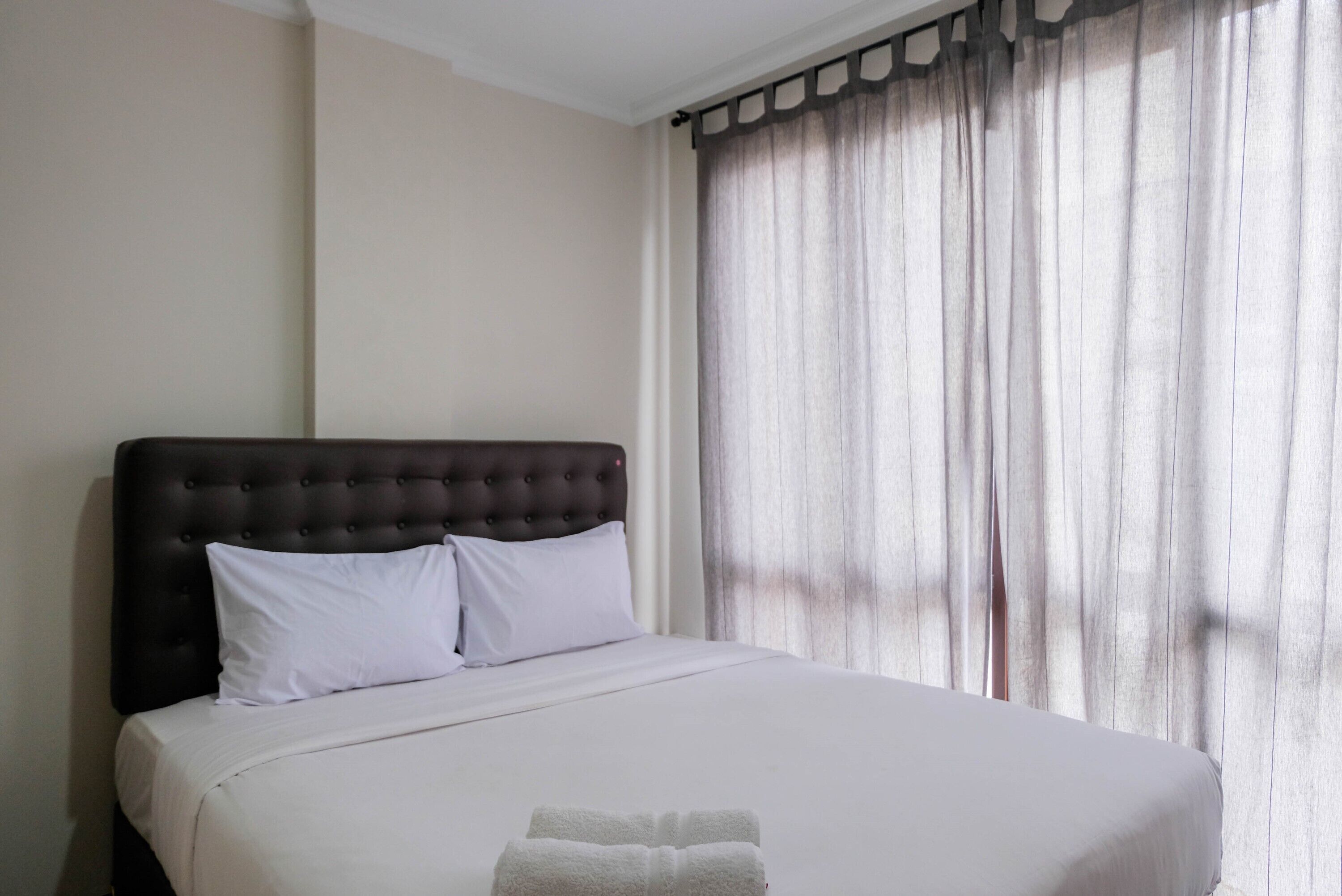 A neatly made bed with a dark headboard and white bedding, a window with sheer curtains, and a white towel on the bed.