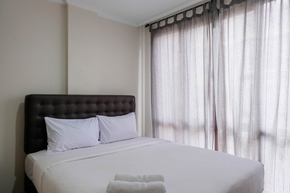A neatly made bed with a dark headboard and white bedding, a window with sheer curtains, and a white towel on the bed.