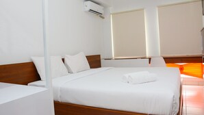 A hotel room with a wooden bed, white bedding, an air conditioning unit, and a window with blinds. - Comfy Studio Room at Patraland Urbano Apartment (Bekasi)