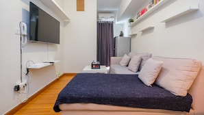 A modern hotel room with a large bed, a flat-screen TV mounted on the wall, a small table with a lamp, and a refrigerator. - Pool View and Homey 2BR Apartment @ Springlake Summarecon (Bekasi)