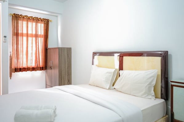 A neatly made bed with white linens and pillows, a wooden wardrobe, and a window with orange curtains.