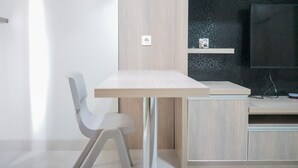 A modern, minimalist room with a wooden desk, a white chair, and a television on a dark patterned wall. - Comfy Studio at Trimezia Beverly Apartment By Travelio (Gading Serpong)