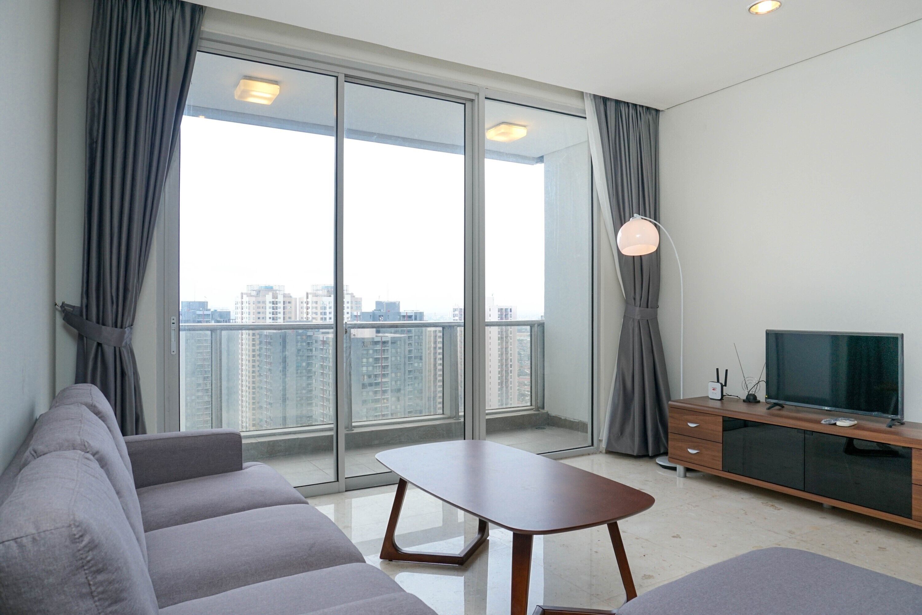Una sala de estar moderna con un sofá, una mesa de centro, un televisor sobre un mueble de madera y un gran ventanal corredero que da a un balcón con vistas a la ciudad.