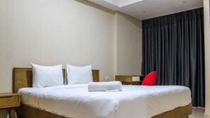 Una cama bien hecha con sábanas blancas y una toalla enrollada, una mesita de noche de madera, una silla roja y una cortina oscura en una habitación de hotel.