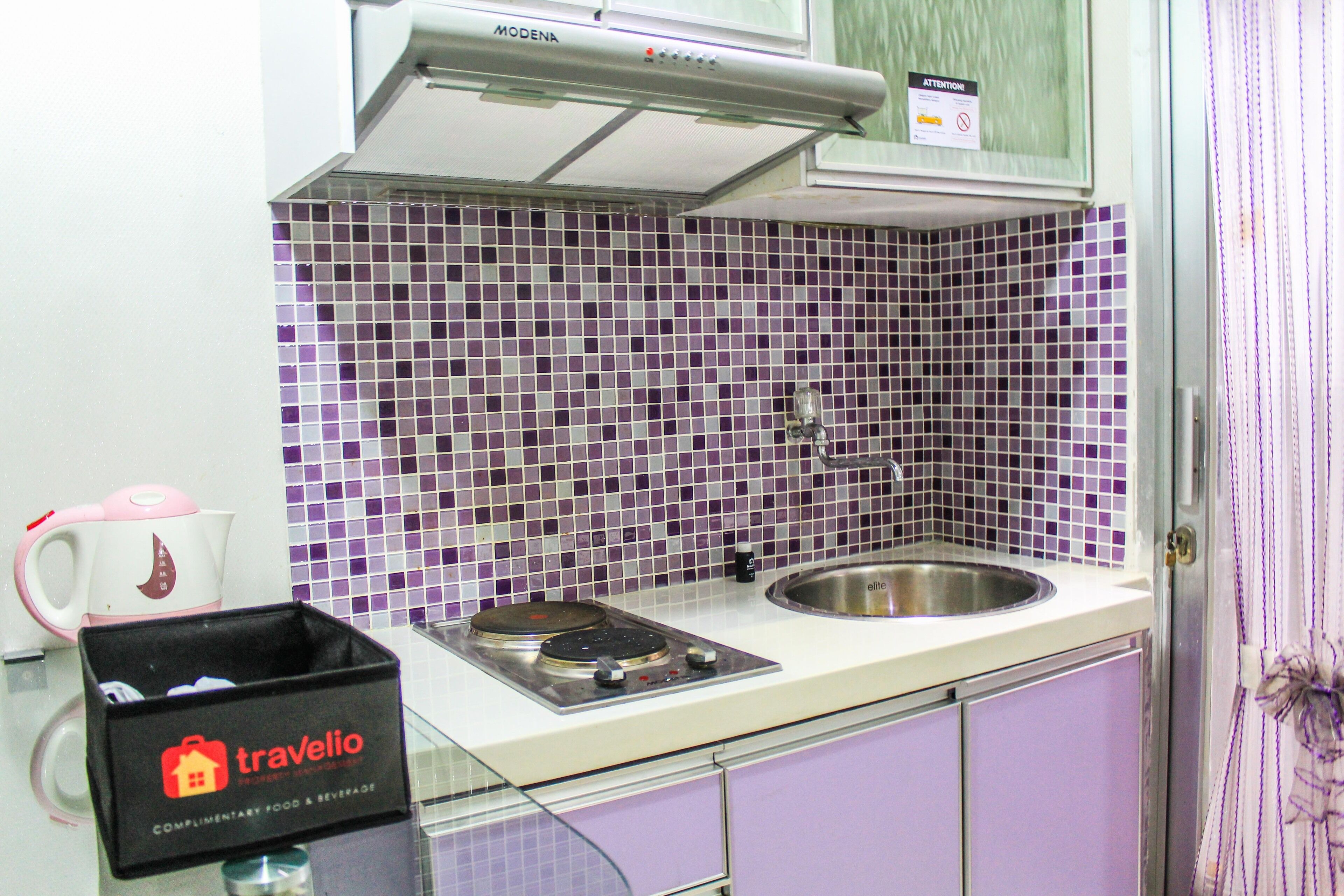 Cocina con salpicadero de azulejos morados, un lavamanos de acero inoxidable y una caja de travelio sobre la encimera.