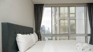 A hotel room with a large bed, a headboard, a nightstand, and a view of the city through a window. - Cozy 2BR at Empyreal Epicentrum Apartment By Travelio (Jakarta)