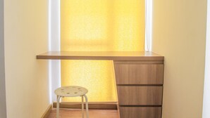 Ein Schreibtisch an der Ecke mit Hocker, ein Fenster mit Jalousien und eine Holzkommode.
