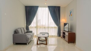 A modern living room with a sofa, a small table, a TV on a wooden stand, and a window with curtains. - Comfy and Spacious 2BR The Masterpiece Epicentrum Apartment By Travelio (Jakarta)
