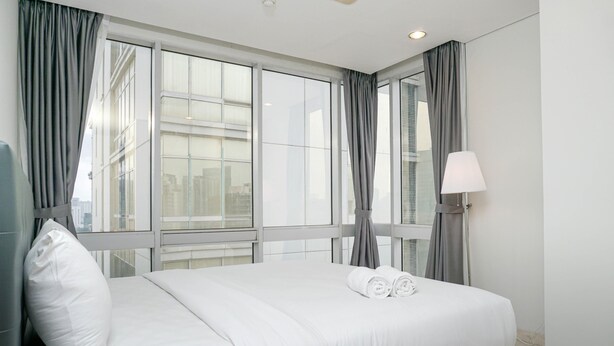A hotel room with a large bed, grey curtains, a white lamp, and a view of the city through large windows. - Comfortable Deluxe 2BR at The Empyreal Condominium Epicentrum Apartment By Travelio (Jakarta)