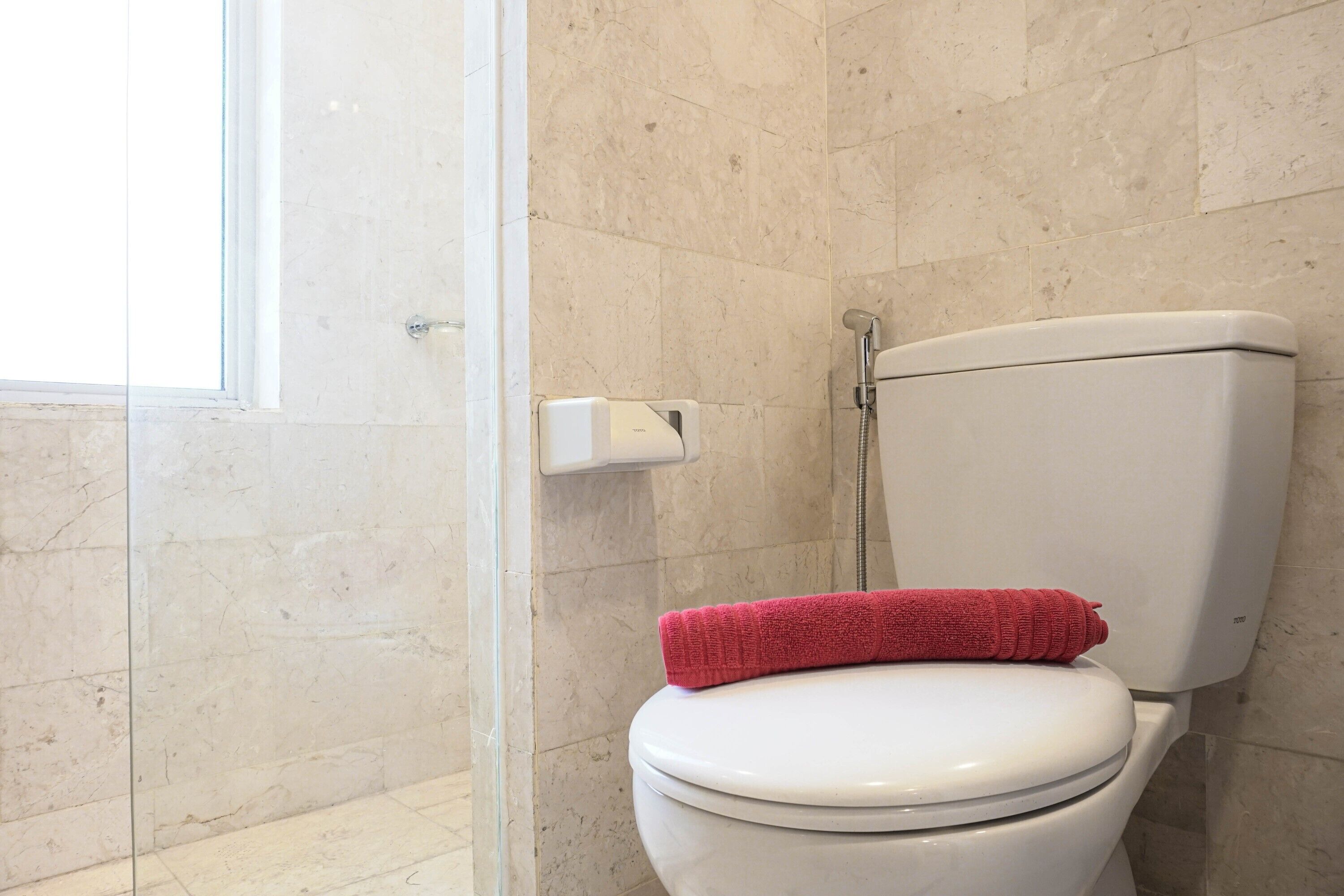 A bathroom with a white toilet, a red towel on the seat, and a shower area with beige tiles.