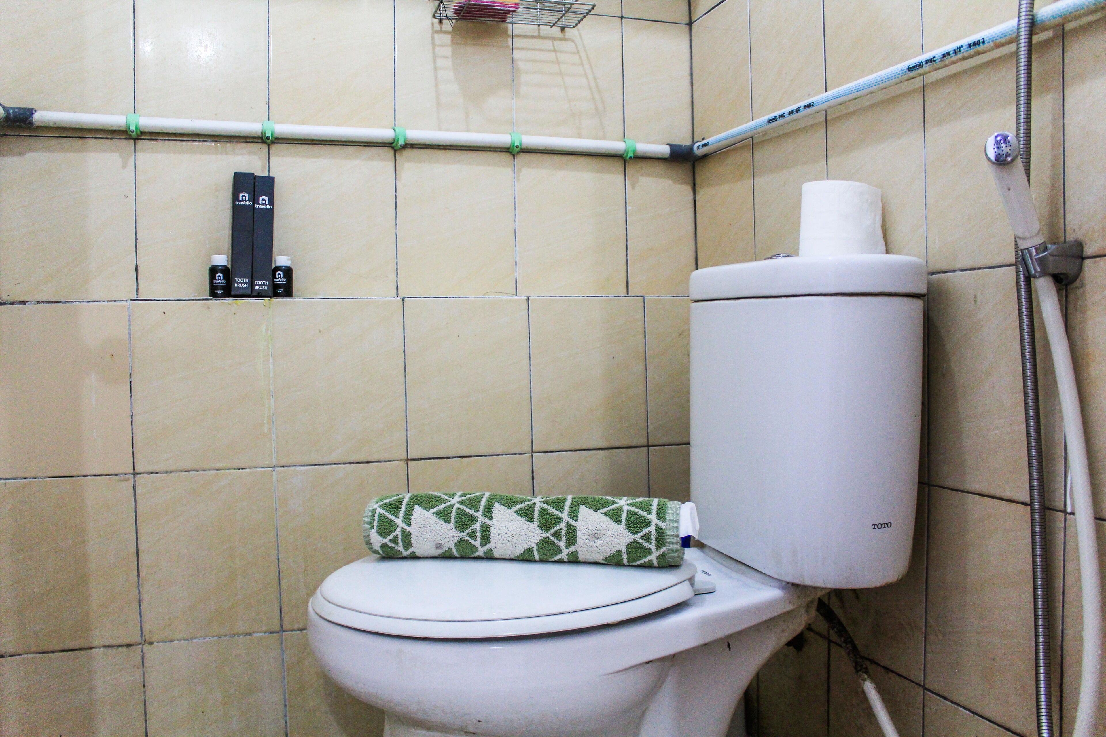 Un inodoro con un dispensador de papel higiénico de diseño verde y blanco, un rollo de papel higiénico blanco y una tubería metálica que recorre la pared.