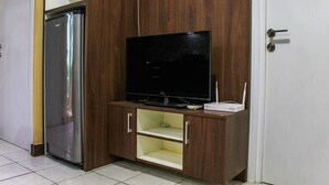 A compact living space with a built-in TV on a wooden cabinet, a small refrigerator, and a door with a handle. - Comfortable and Tranquil 2BR Apartment at Green Pramuka By Travelio (Jakarta)