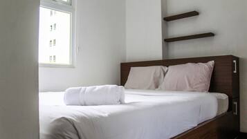 Una cama bien hecha con ropa blanca y una toalla blanca enrollada, una ventana con vista a edificios y un estante montado en la pared.