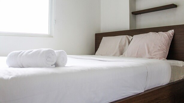 A neatly made bed with white linens and a rolled towel, a wooden headboard, and built-in shelves above the bed. - Comfortable and Tranquil 2BR Apartment at Green Pramuka By Travelio (Jakarta)