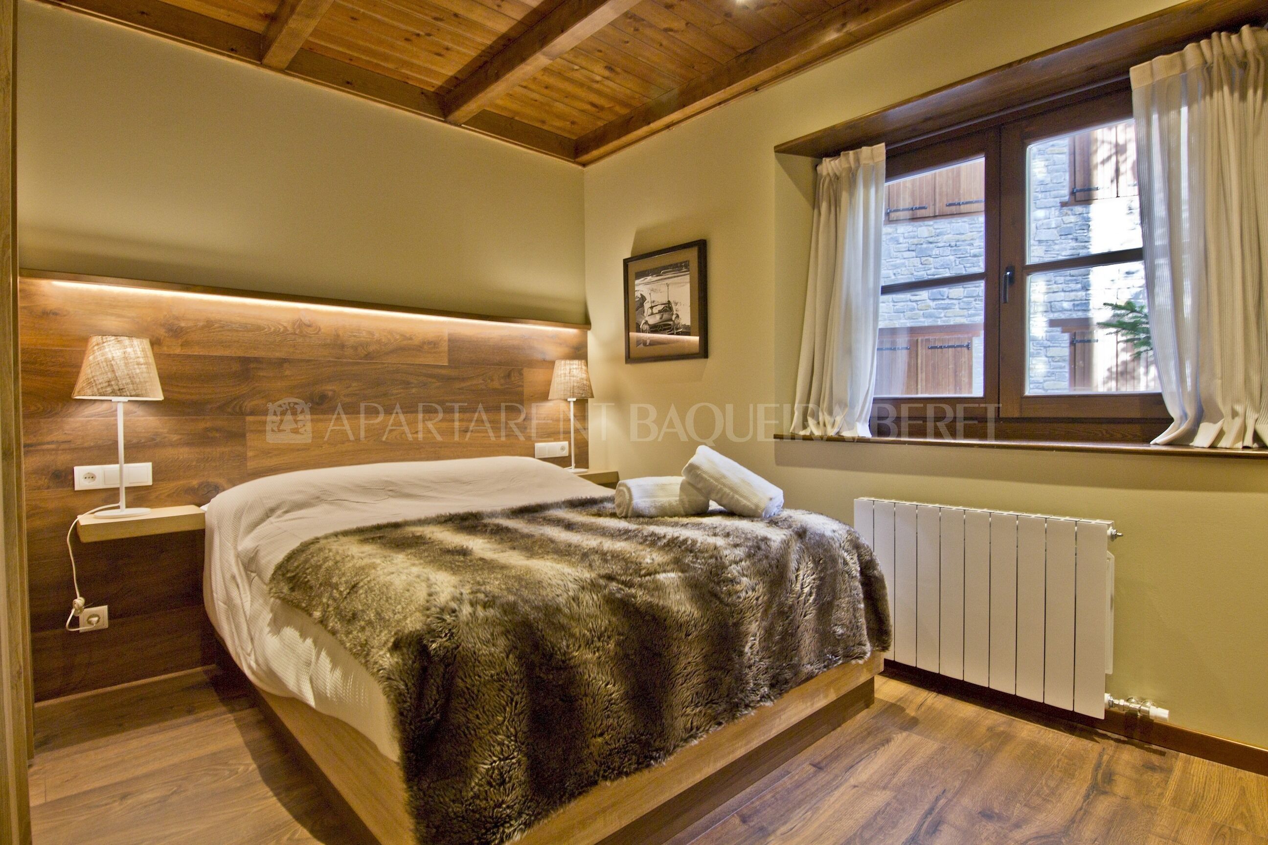 A bedroom with a wooden headboard, a bed with a fluffy blanket, a nightstand with a lamp, and a window with curtains.