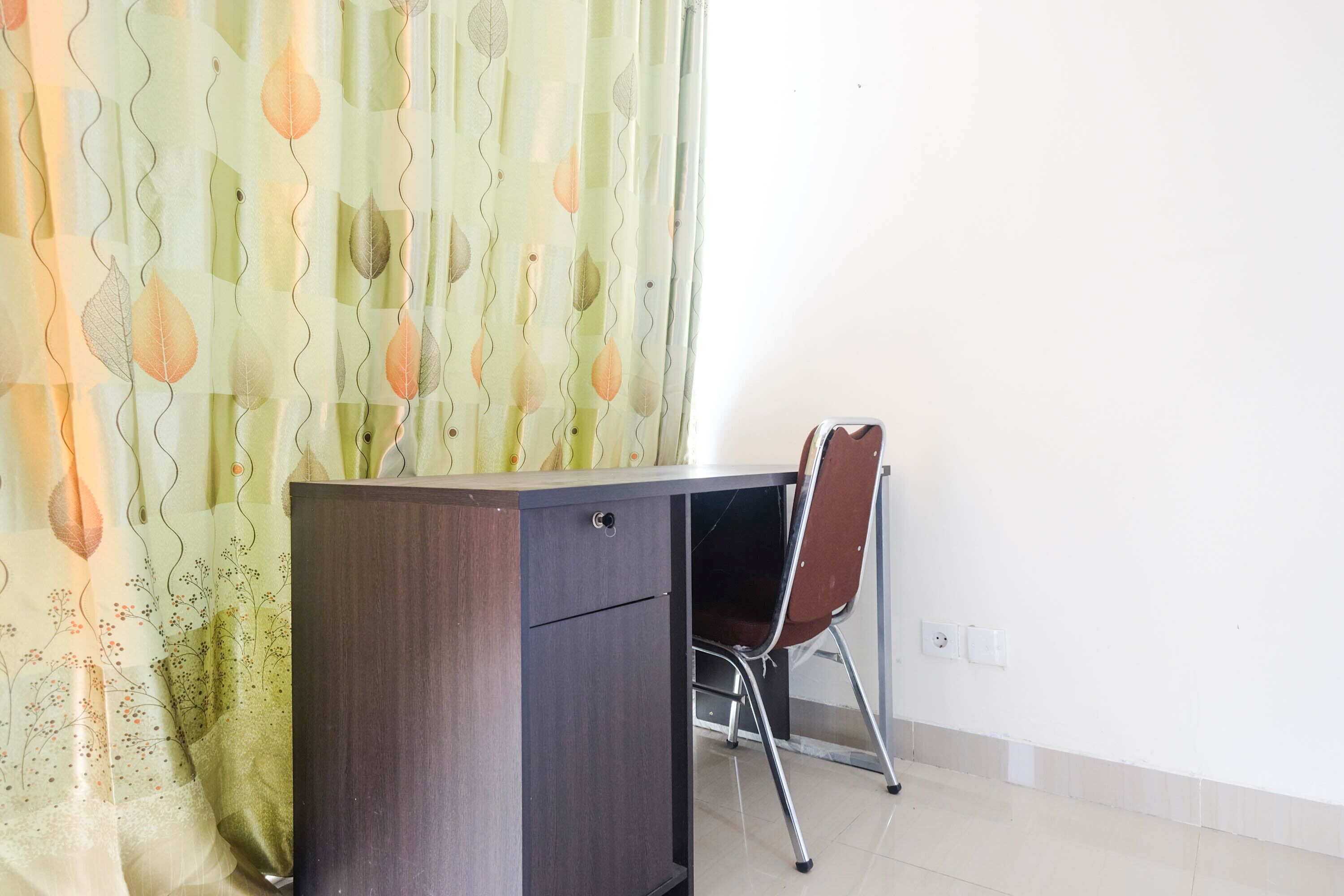 A room with a wooden desk and a chair, a patterned curtain, and a plain white wall.