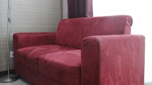 A red sofa in a room with a floor lamp and a window with curtains. - Prime & Comfy Studio @ Tamansari La Grande Apartment By Travelio (Bandung)