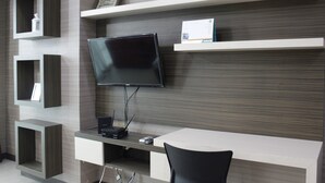 A modern office space with a flat-screen TV mounted on a wooden wall, a desk with a chair, and built-in shelving units. - Prime & Comfy Studio @ Tamansari La Grande Apartment By Travelio (Bandung)