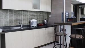 A modern kitchen with a black countertop, white cabinets, and a tiled backsplash. There are two bar stools and a coffee machine on the counter. - Prime & Comfy Studio @ Tamansari La Grande Apartment By Travelio (Bandung)