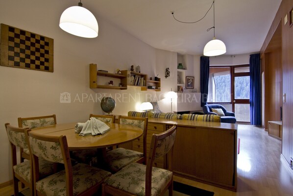 Un comedor con una mesa redonda y sillas estampadas, una cocina con armarios de madera y una silla azul, y vistas del exterior a través de una ventana.