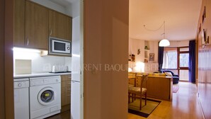 A compact kitchen with a microwave, washing machine, and wooden cabinets. A dining area with a sofa and table is visible in the background.