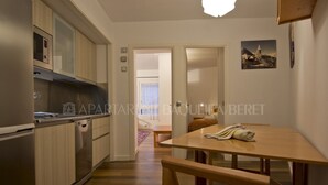 A modern kitchen with built-in appliances, a dining area with a wooden table and bench, and a view of the living room through an open door. - Urogallo - Edificio Aneto (Baqueira)