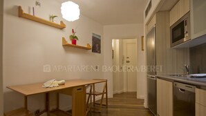 A modern kitchen with wooden cabinets, a built-in microwave, and a stainless steel refrigerator. There is a wooden table with a chair and a small shelf with plants. - Urogallo - Edificio Aneto (Baqueira)
