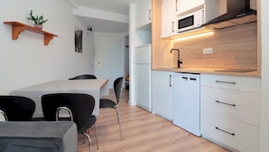 Une cuisine moderne avec des meubles blancs, un four à micro-ondes et un lave-vaisselle. On y trouve une table à manger avec des chaises, ainsi qu’une plante en pot posée sur une étagère en bois.