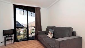 Un salon moderne avec un canapé gris, un petit meuble noir pour la télévision et un balcon offrant une vue sur les montagnes.