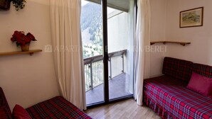 A room with a bed, a sofa, a balcony with a view of a forested mountain, and a window with curtains. - Garona - Edificio Aneto (Baqueira)