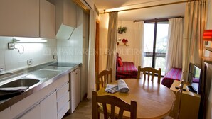 A compact hotel room with a kitchenette, a dining area with a table and chairs, a bed with a red and black checkered blanket, and a TV on a stand. - Garona - Edificio Aneto (Baqueira)