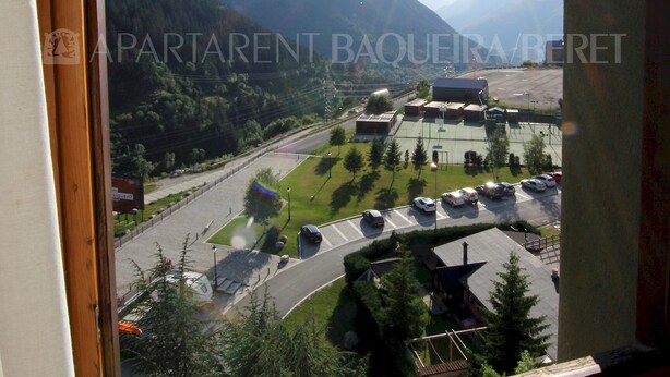 A view from a window of a building with a mountain landscape, a parking lot, and a few buildings in the distance. - Craba - Edificio Aneto (Baqueira)