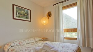 A bedroom with a floral bedspread, a bedside lamp, a framed picture on the wall, and a window with curtains. - Craba - Edificio Aneto (Baqueira)