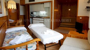 A hotel room with two single beds, a wooden chair, a small table, a TV, and a kitchenette in the background. - Craba - Edificio Aneto (Baqueira)