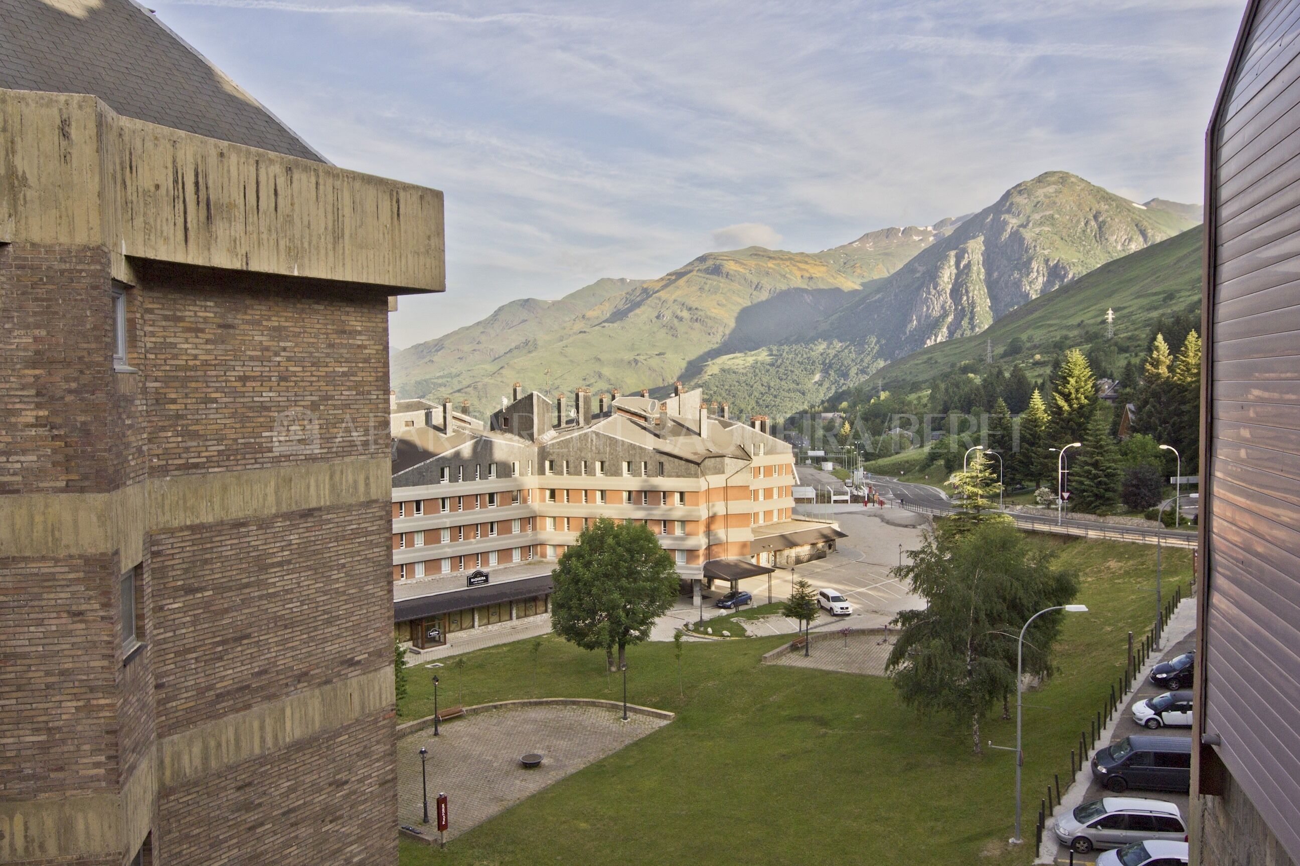 A multi-story building with a brick facade, a parking area with cars, and a view of mountains in the background.