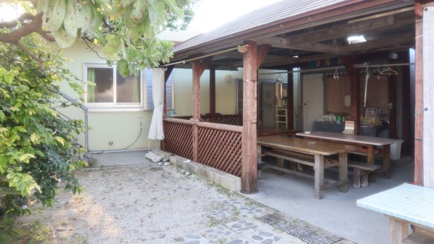 A covered outdoor seating area with wooden tables and benches, a small garden with a tree, and a building with a sign that reads "ONN". - Minshuku Robinson (Zamami)