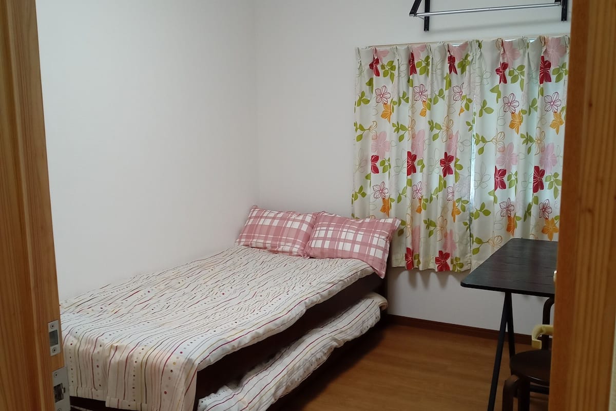 A small, single-bed room with a wooden floor, a small table, and a window with floral curtains.