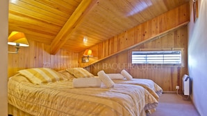 Una habitación con paredes de madera panelada, techo inclinado, dos camas con ropa de cama a rayas, una ventana con persianas y un calentador fijado a la pared.
