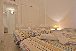 Une chambre avec deux lits, une armoire, une table de chevet avec une lampe, et une salle de bain visible à travers une porte ouverte.