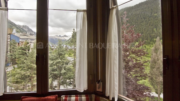 A room with a large window offering a view of a mountainous landscape and a town. - Irissa - Edificio Campalias (Baqueira)