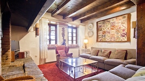 Een gezellige woonkamer met een stenen open haard, een rood tapijt, een glazen salontafel en een groot, geborduurd wandkleed.