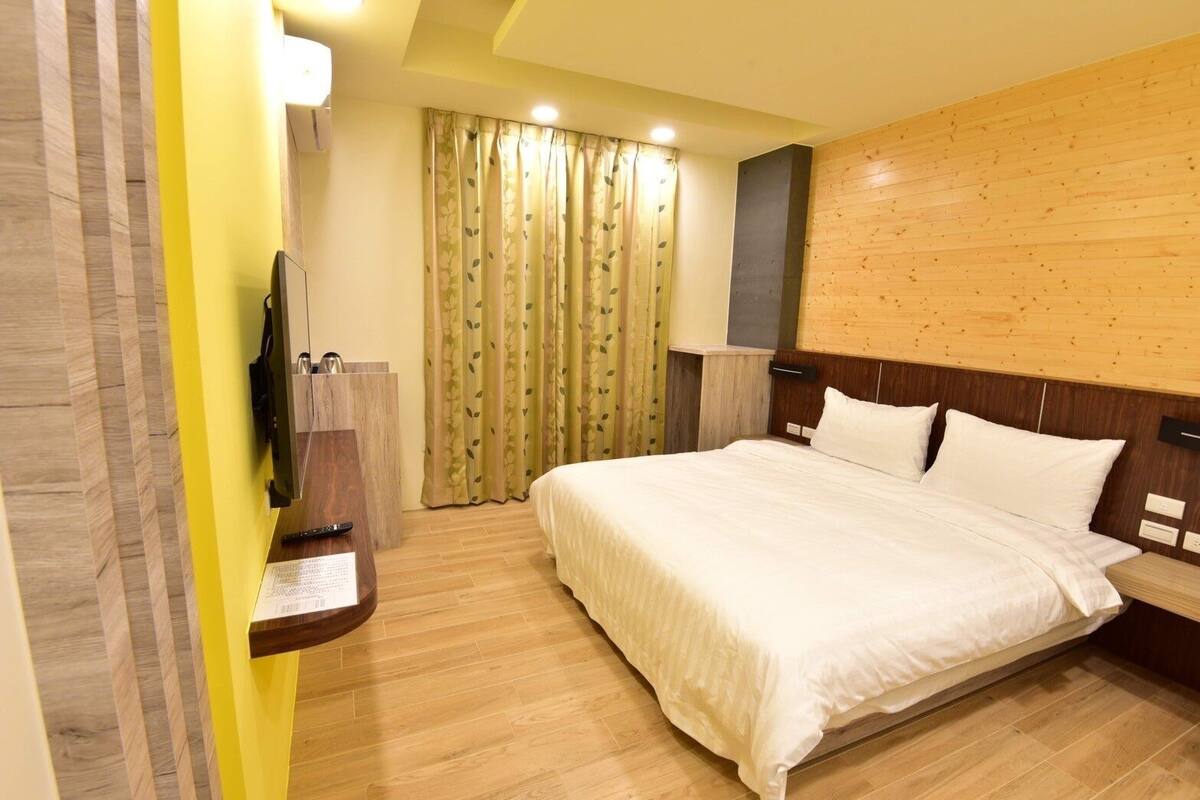 A hotel room with a wooden headboard, a bed with white bedding, a wooden nightstand, a flat-screen TV mounted on the wall, and a window with floral curtains.