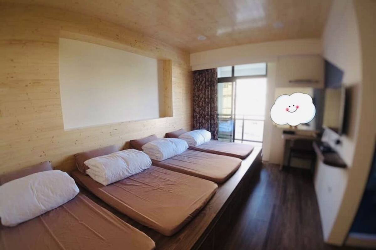 A hotel room with a long row of beds, a wooden wall, a window with curtains, and a television on a stand.