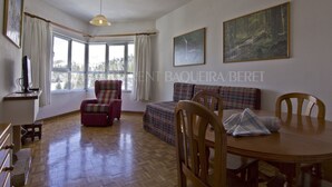 A spacious room with a wooden floor, a dining table with chairs, a sofa, a red armchair, and two paintings on the wall.