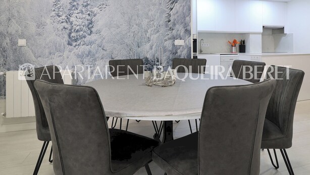 Un comedor moderno con una mesa redonda y sillas tapizadas en gris, una cocina al fondo y un mural en la pared que representa un bosque nevado.