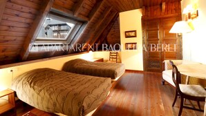 A room with two beds, wooden flooring, a dining table with chairs, a window with a view, and a door with a decorative panel. - Cumbres - Nin de Beret (Baqueira)