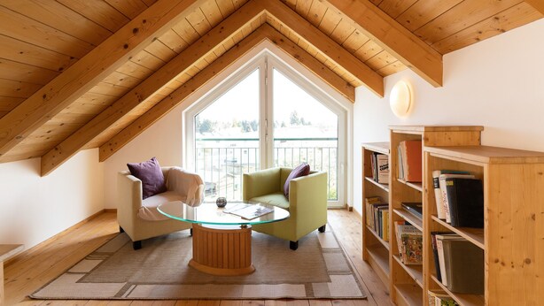 Un acogedor dormitorio en el ático con techo de madera, una mesa de centro de vidrio, dos sillones, una estantería y un gran ventanal con vistas.