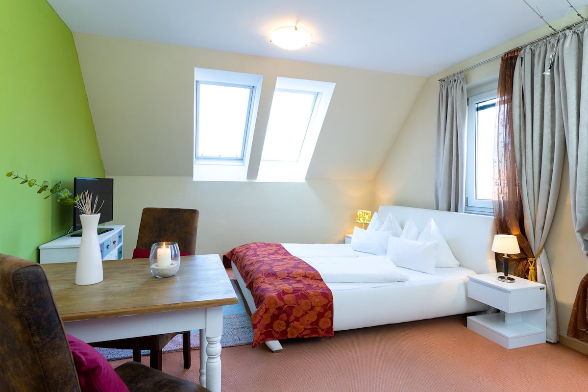 A bedroom with a bed, bedside table, lamp, and a small desk with a plant and vase.