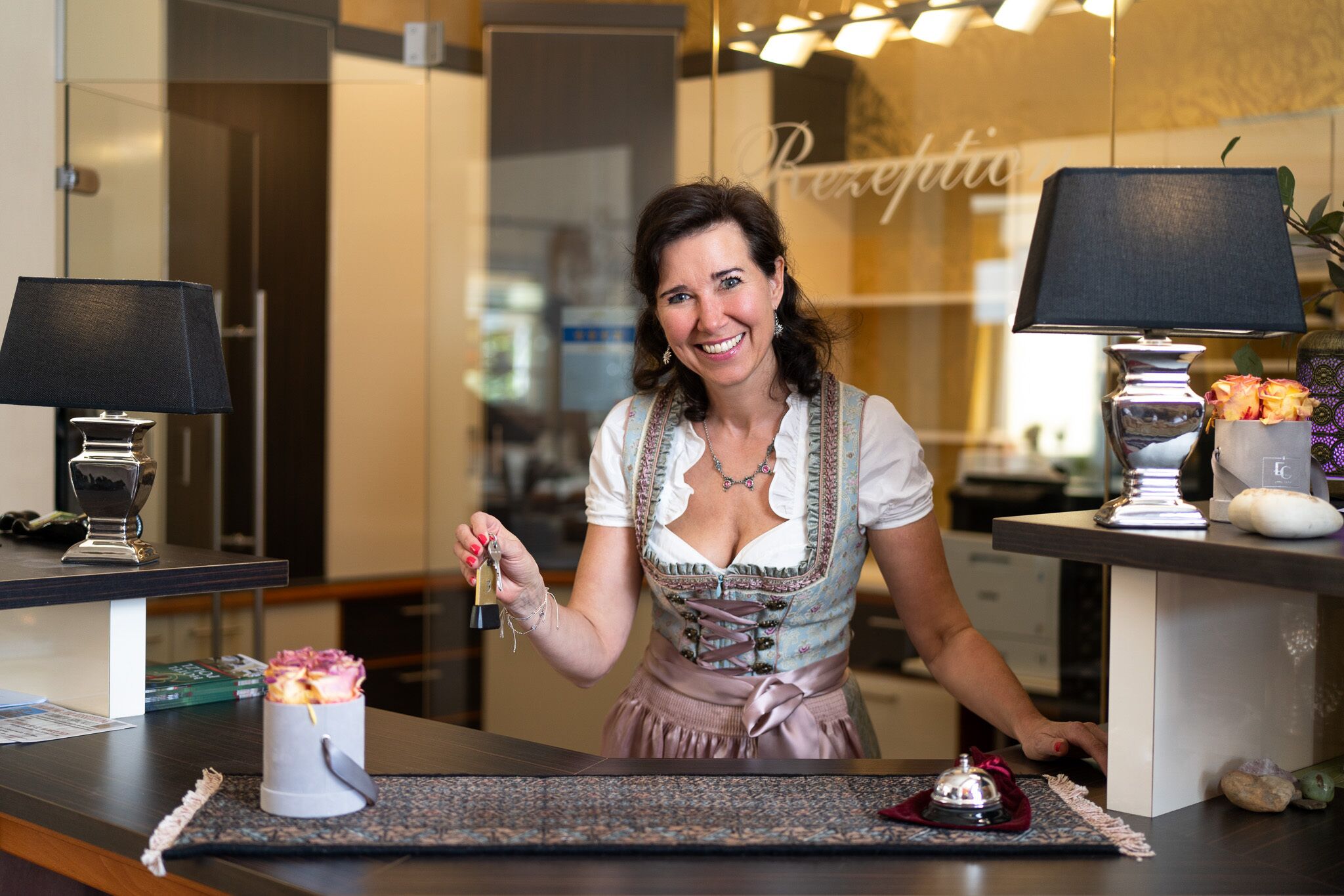 Une femme vêtue d’un costume traditionnel, tenant une clé, se tient dans le hall d’un hôtel moderne, où l’on aperçoit un comptoir d’accueil, des lampes et un vase décoratif.