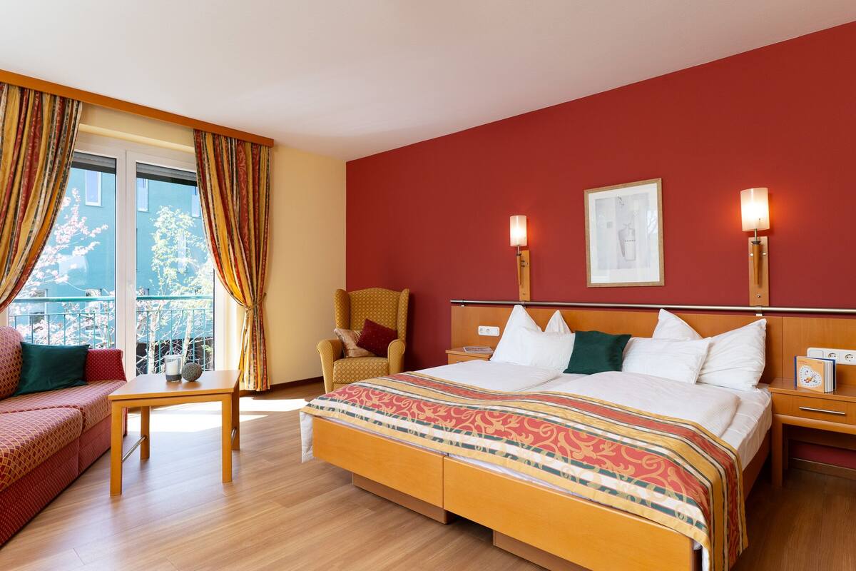 A hotel room with a large bed, a wooden nightstand, a chair, a small table, and a balcony with curtains.